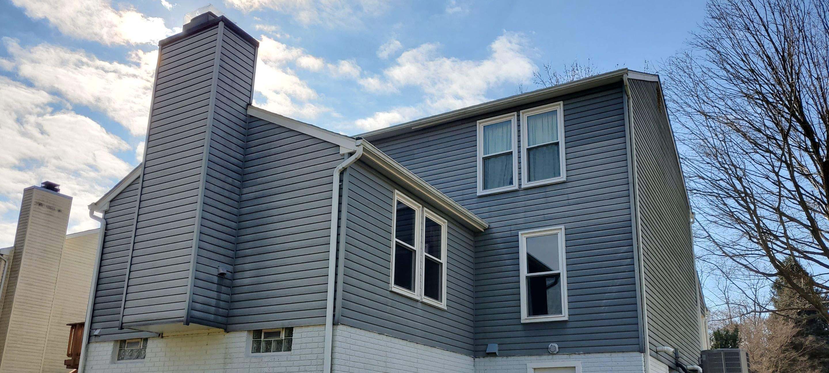 Two-story blue-gray house with white trim and multiple windows, brick base, under a partly cloudy sky.
