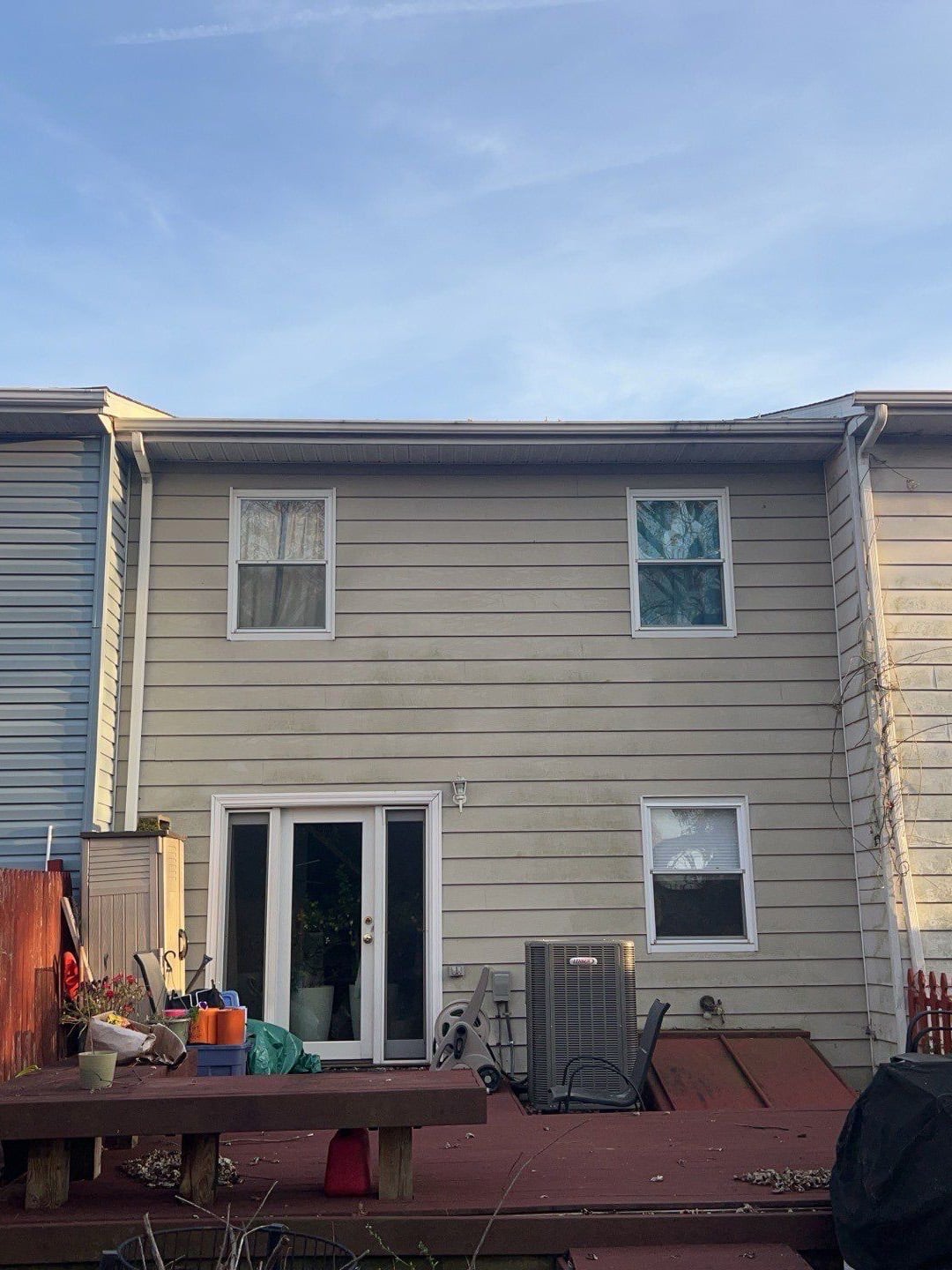 Back of a two-story beige house with a deck, sliding glass doors, two upper windows, and cluttered outdoor items on the deck near an air conditioner unit.