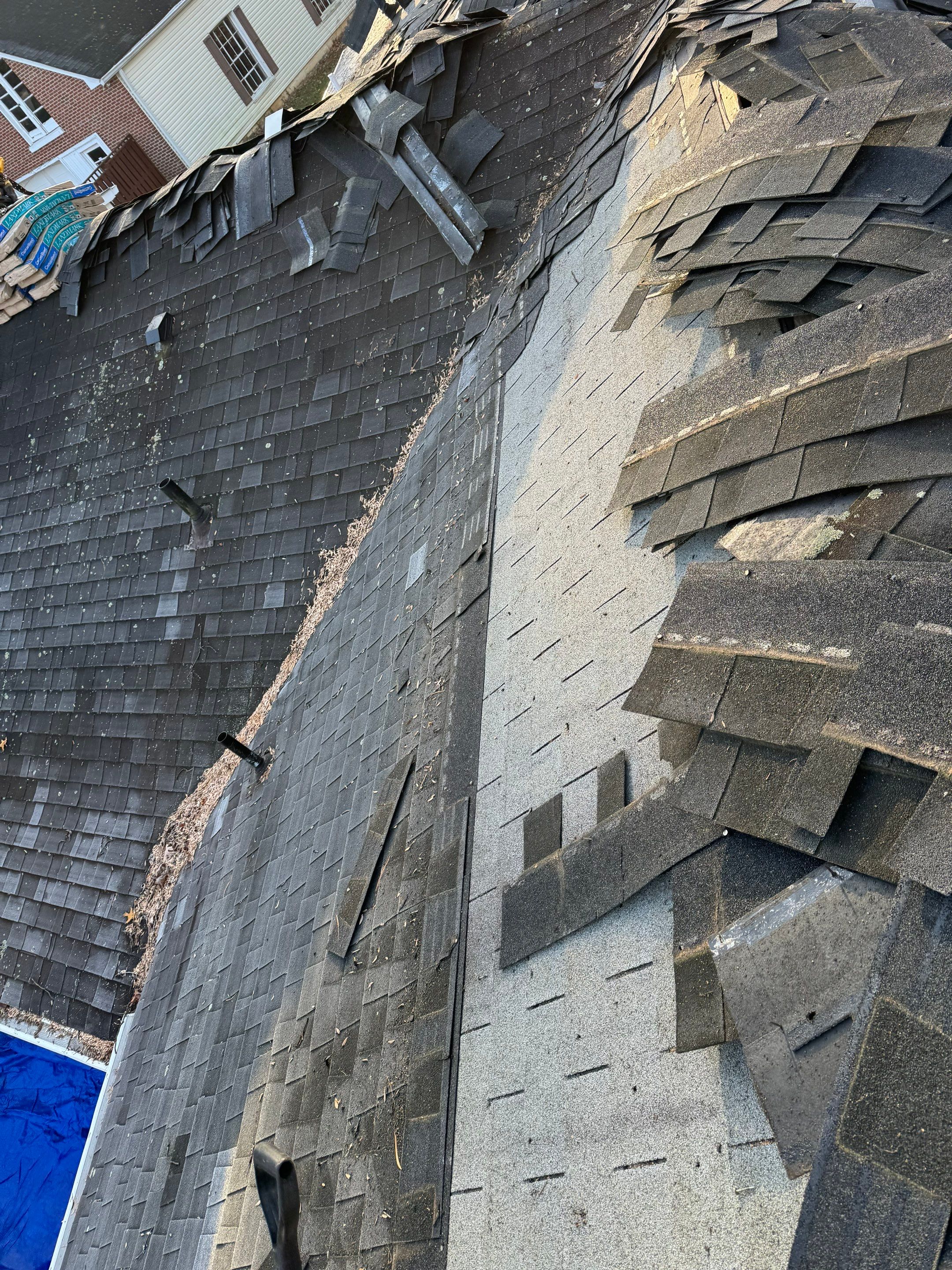 Damaged asphalt shingle roof with loose shingles and stacks of roofing material at the edge; neighboring houses in the background.