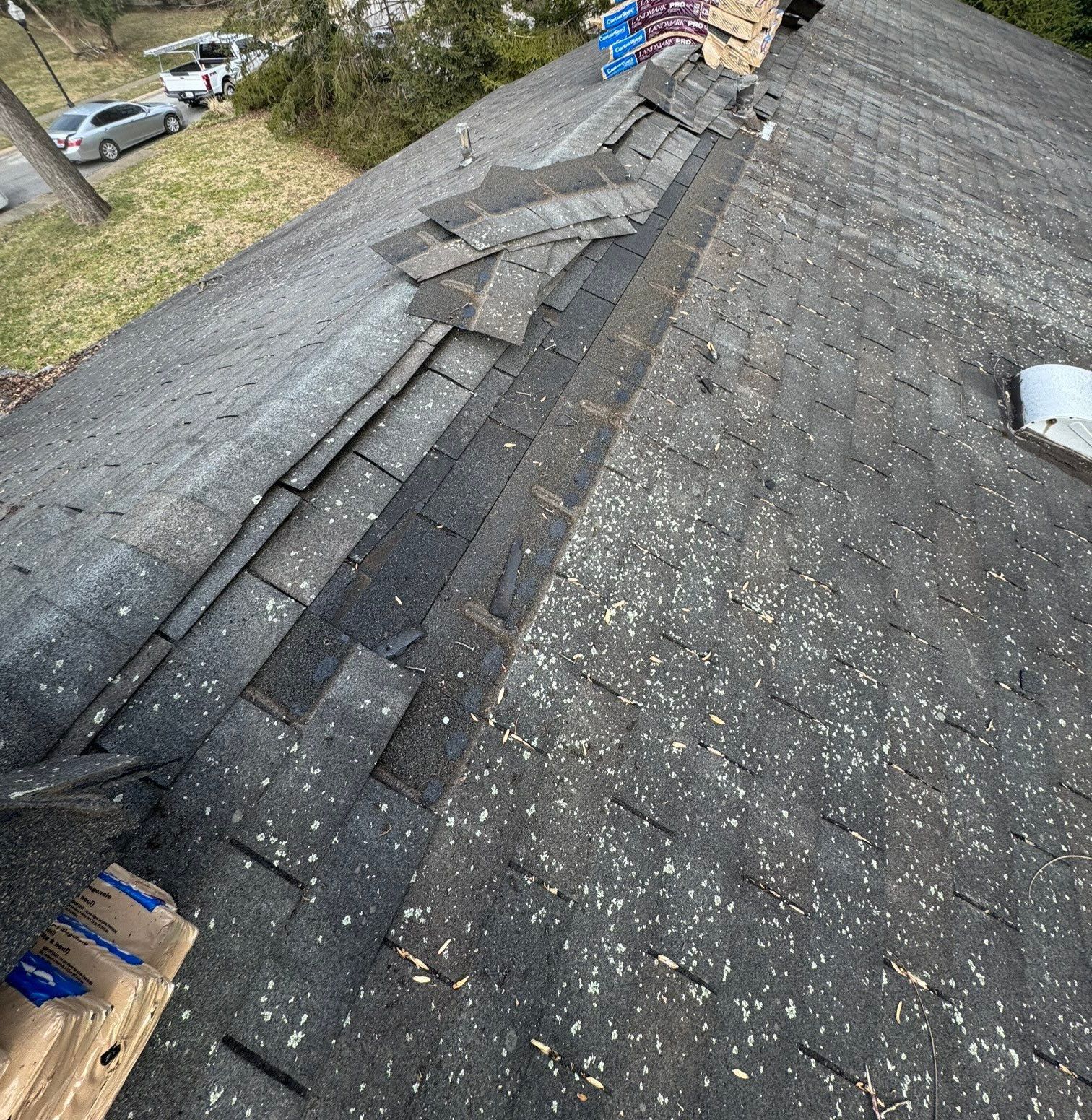 Pitched asphalt shingle roof with dislodged shingles along the ridge and a stack of replacement shingles at the peak, ready for repair