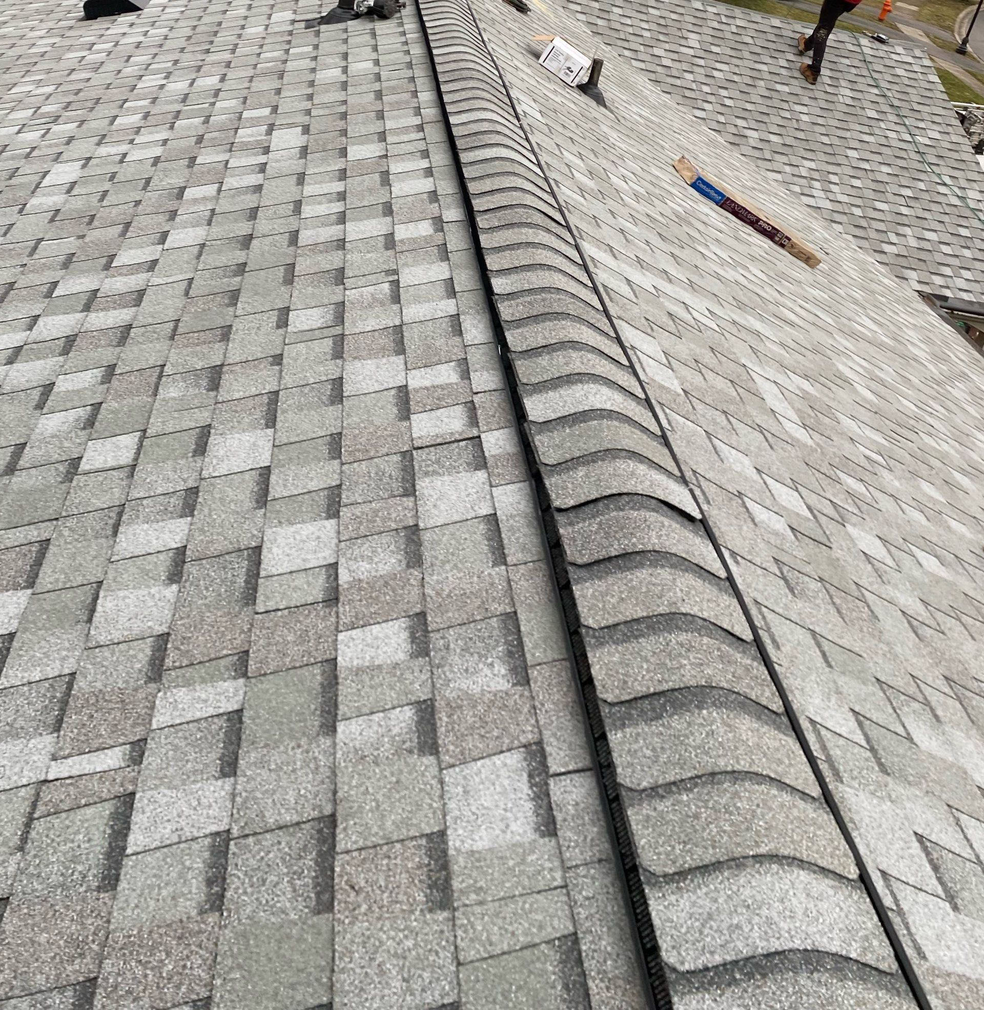 Overhead view of a gray asphalt shingle roof under construction, with a curved ridge vent running along the center and a worker visible at the top edge of the roof.