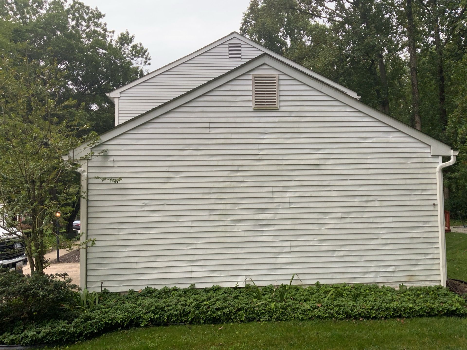 White siding house wall with two attic vents, framed by trees and shrubs in a green yard.