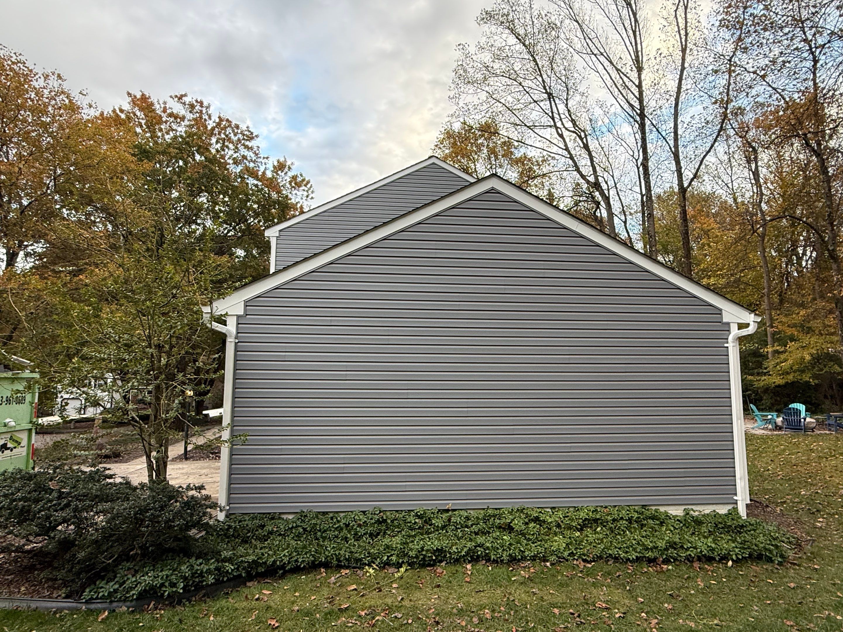 Back of a gray siding house with a triangular roof, surrounded by autumn trees and a green lawn.