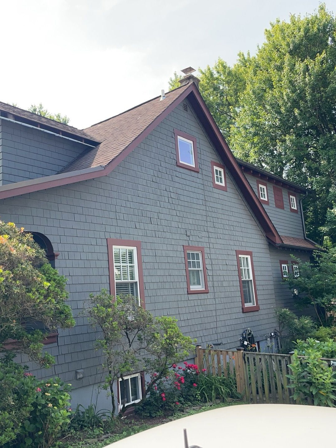 Grey-blue house with red window trim and a steep gable roof, surrounded by greenery and a wooden fence.