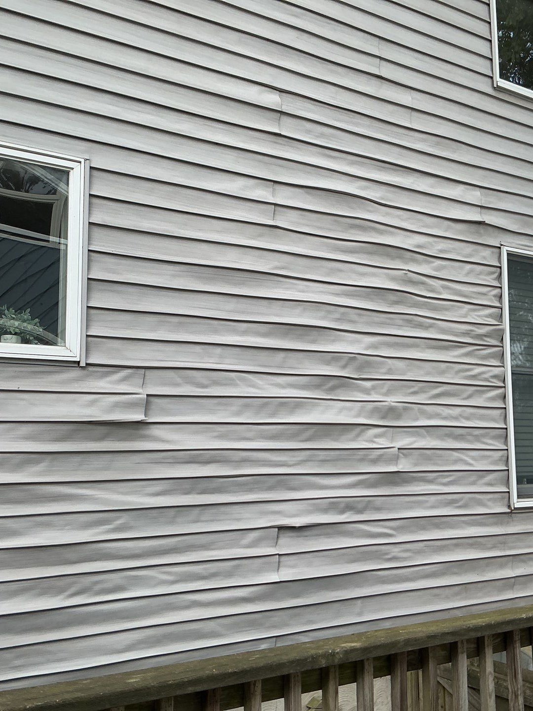 Exterior of a house with light gray horizontal vinyl siding that is buckling and loose in several spots, with windows on the left and right and a wooden railing at the bottom.