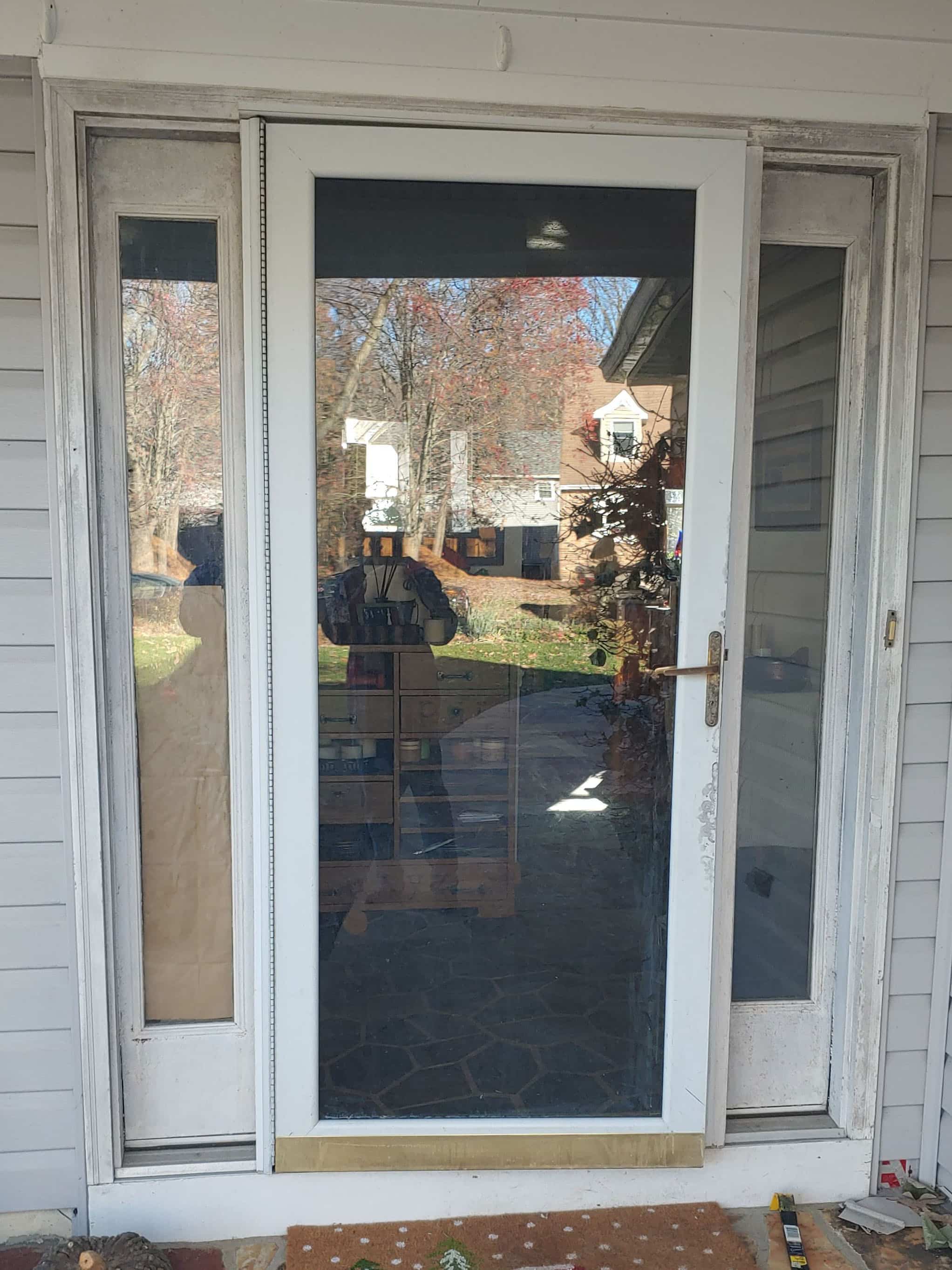 Front entry door with large glass panel and narrow side windows, white frame, reflecting a suburban yard and houses outside.