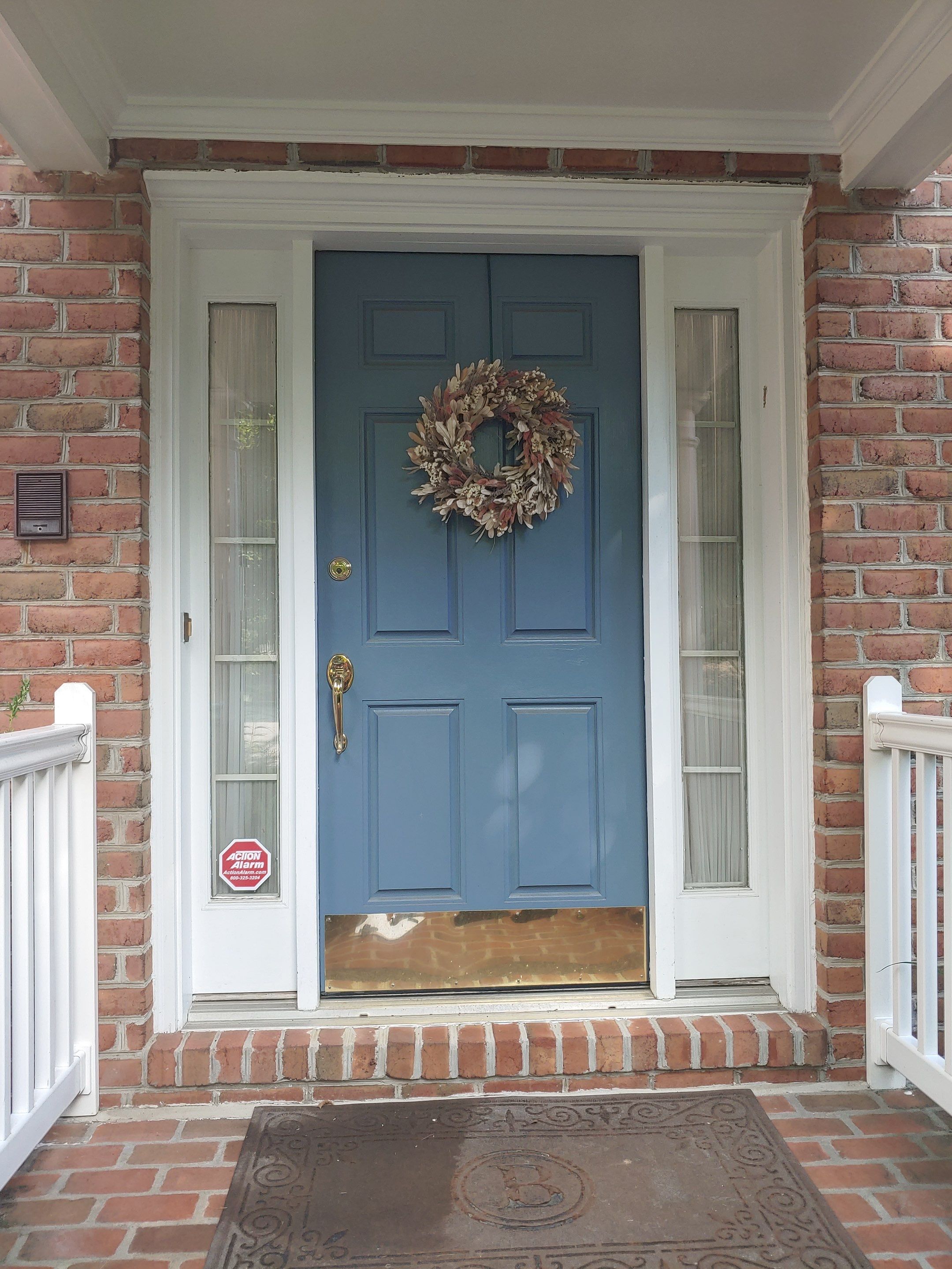 Blue front door with a beige-toned wreath, brick exterior and white trim, flanked by narrow sidelights and brass hardware, entry mat in front.