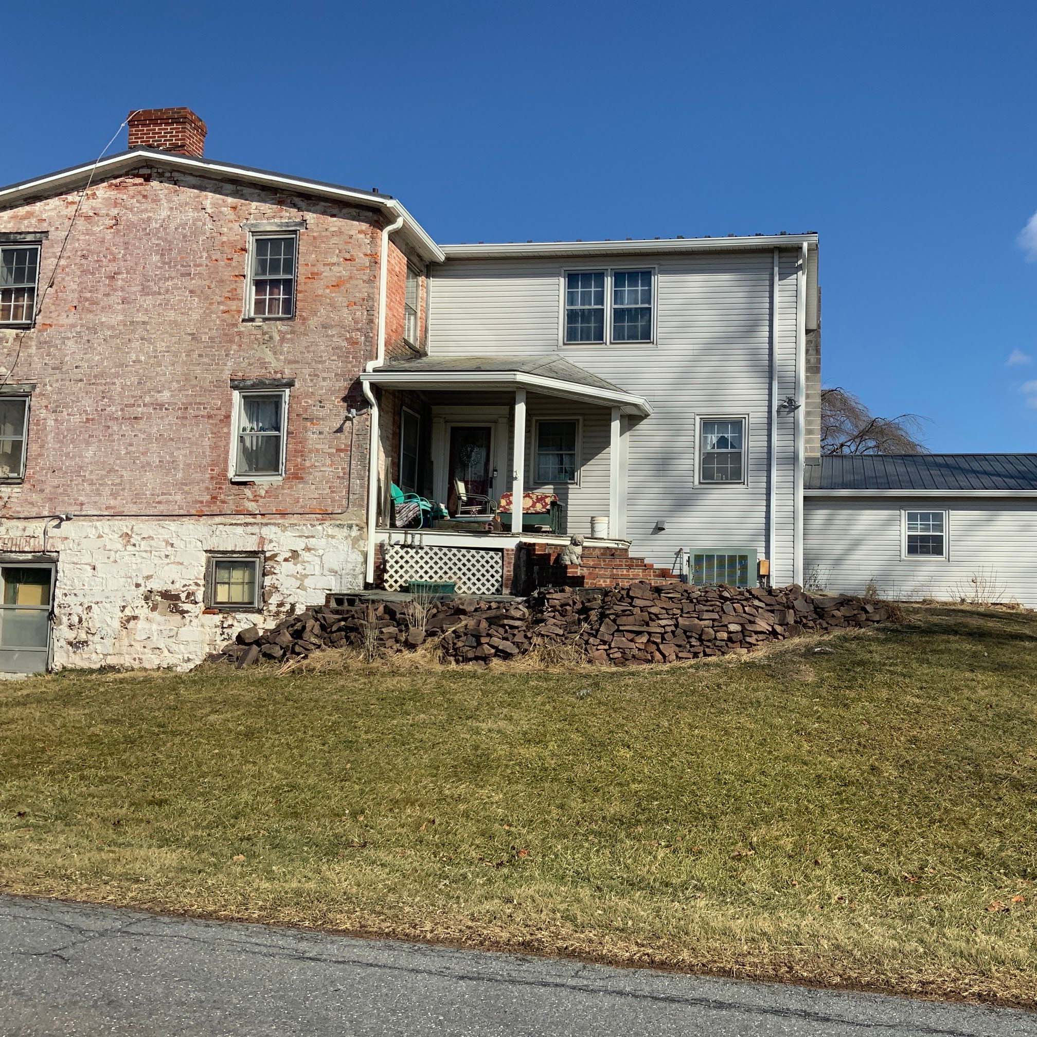 Brick and siding house facade under a blue sky; a rock pile and bare yard in the foreground.
