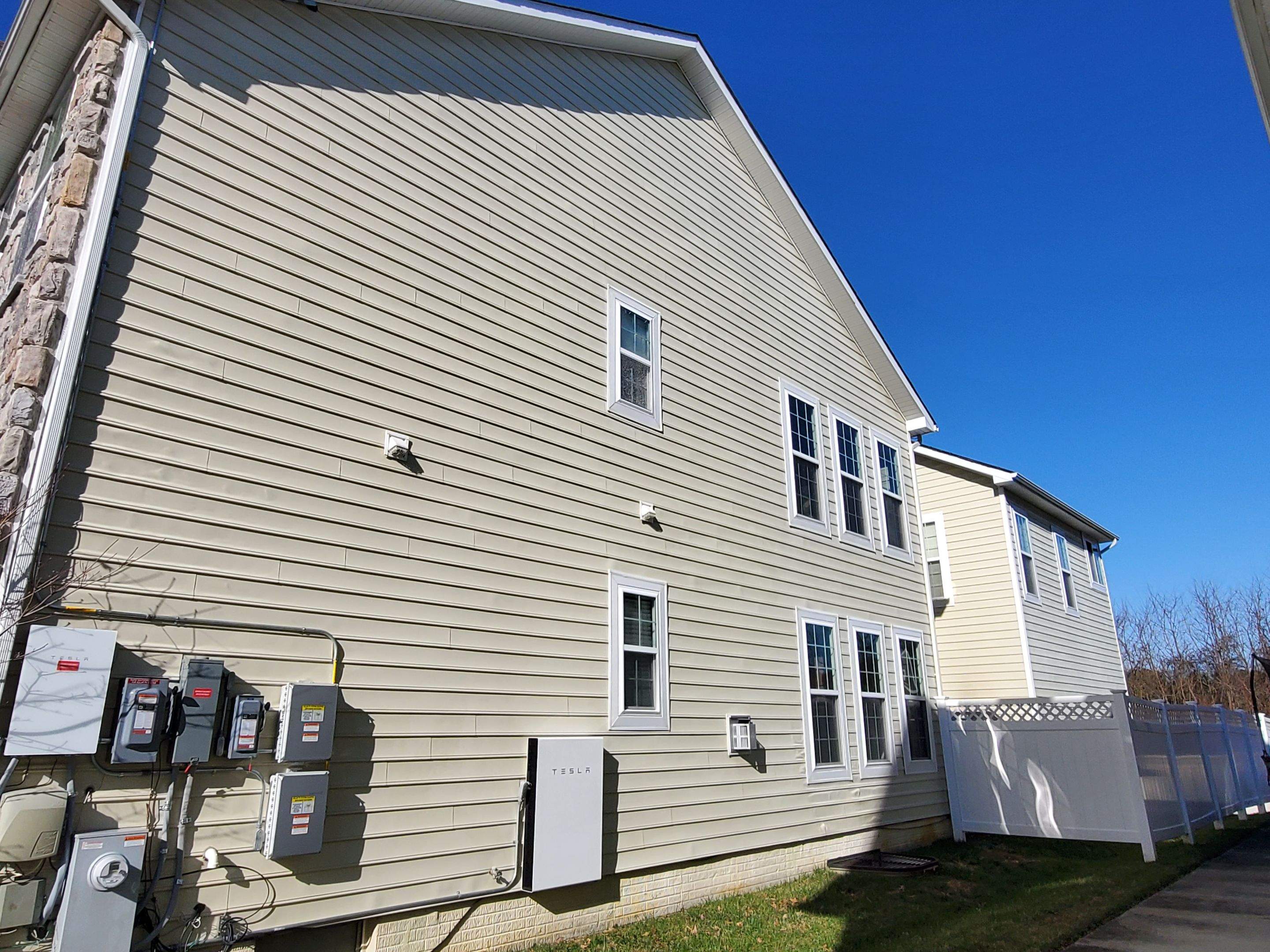 Beige siding exterior wall of a house with white-framed windows and electrical panels mounted along the wall, under a clear blue sky.
