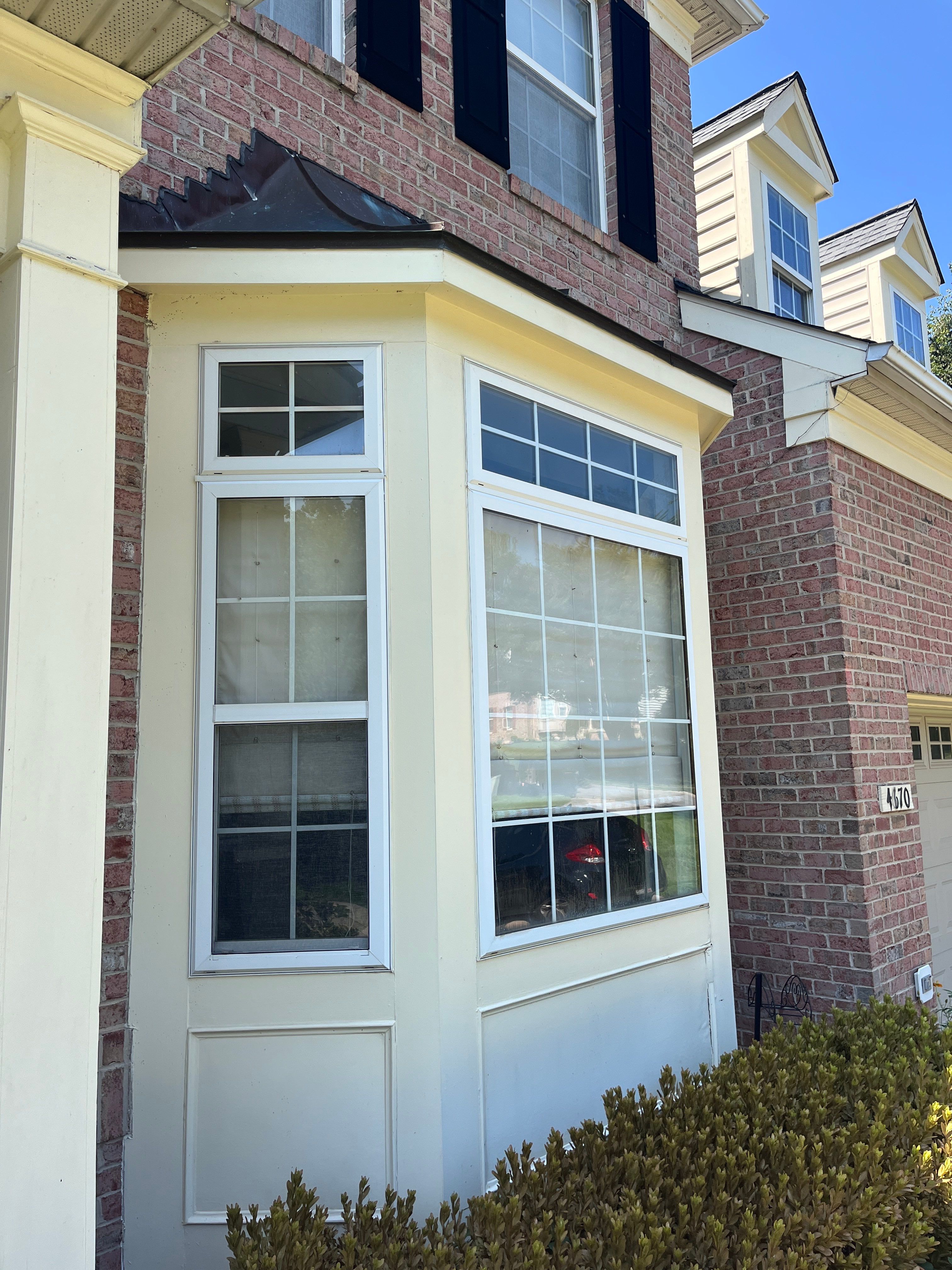 Brick house facade with a large bay window framed in cream trim, black shutters, and trimmed hedges in the foreground.