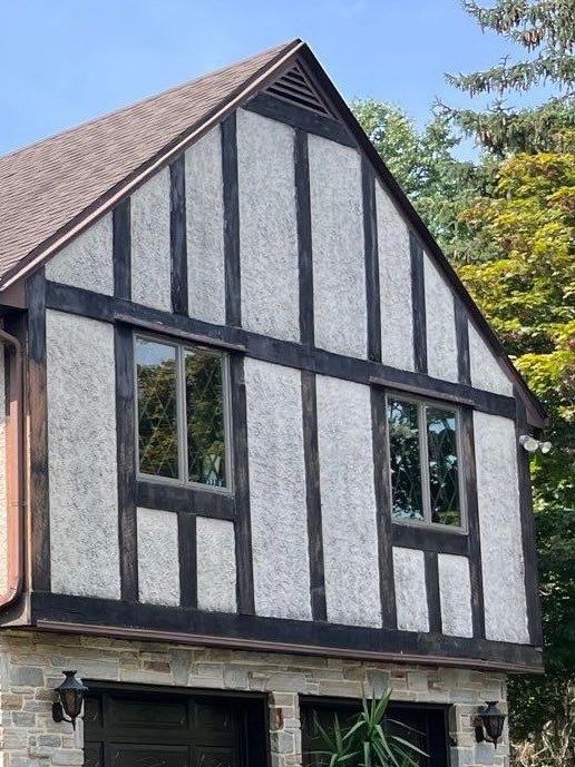 Tudor-style stone-and-timber house facade with gray plaster infill and black wood framing, two windows on upper floor.