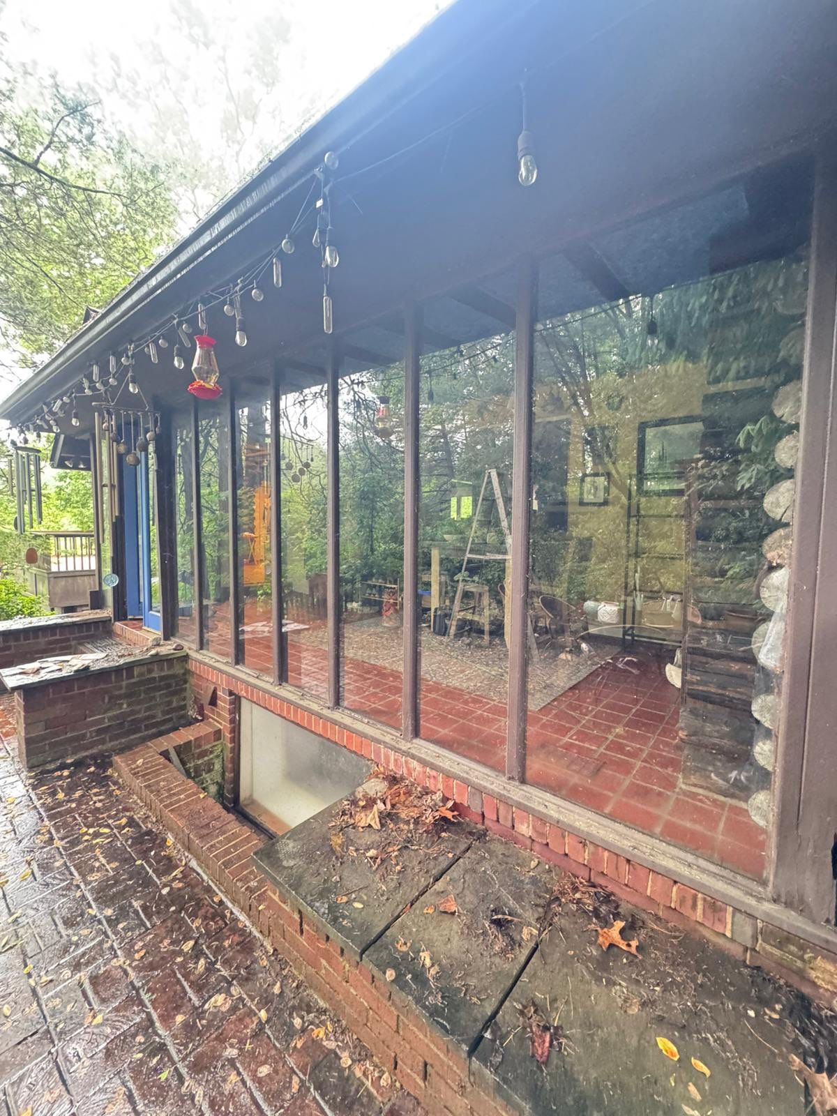 Glass-walled sunroom or porch with string lights along the roof and a brick patio outside, autumn leaves scattered on the ground.