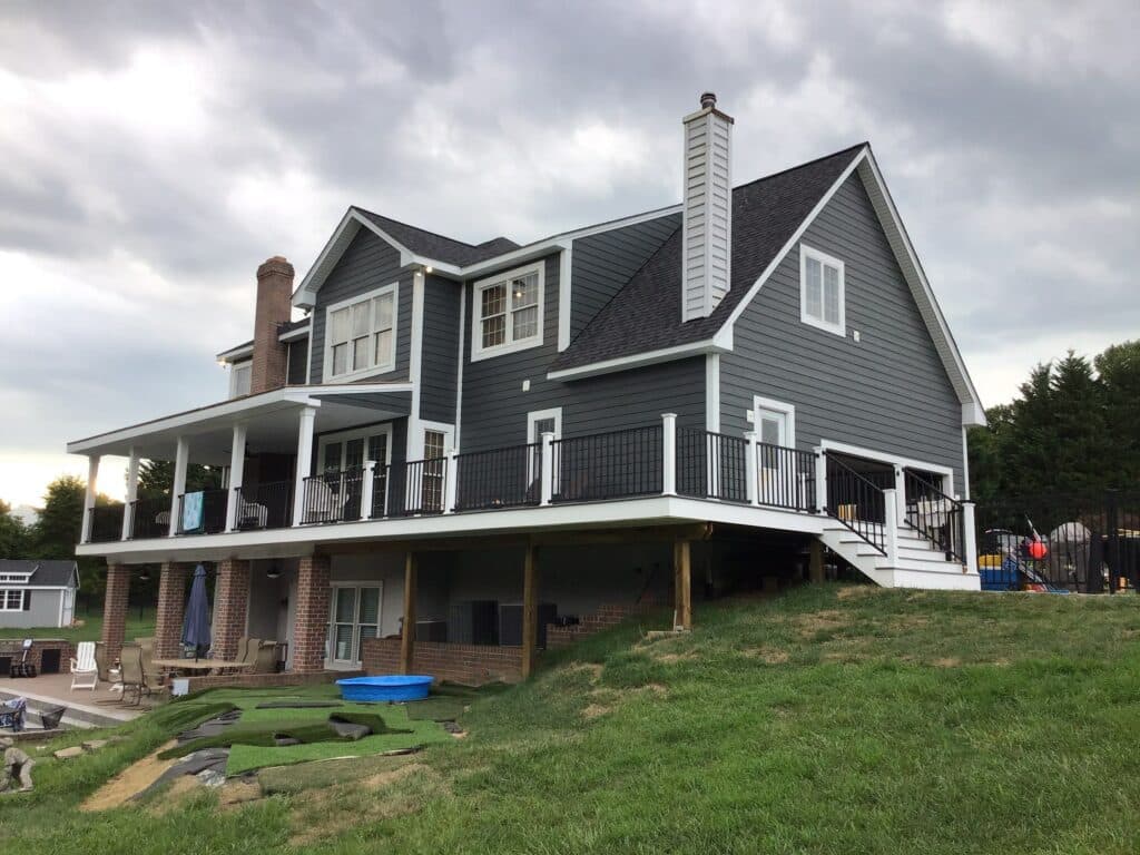 Two-story gray house with white trim on a raised deck and stairs, set against a cloudy sky and green lawn.