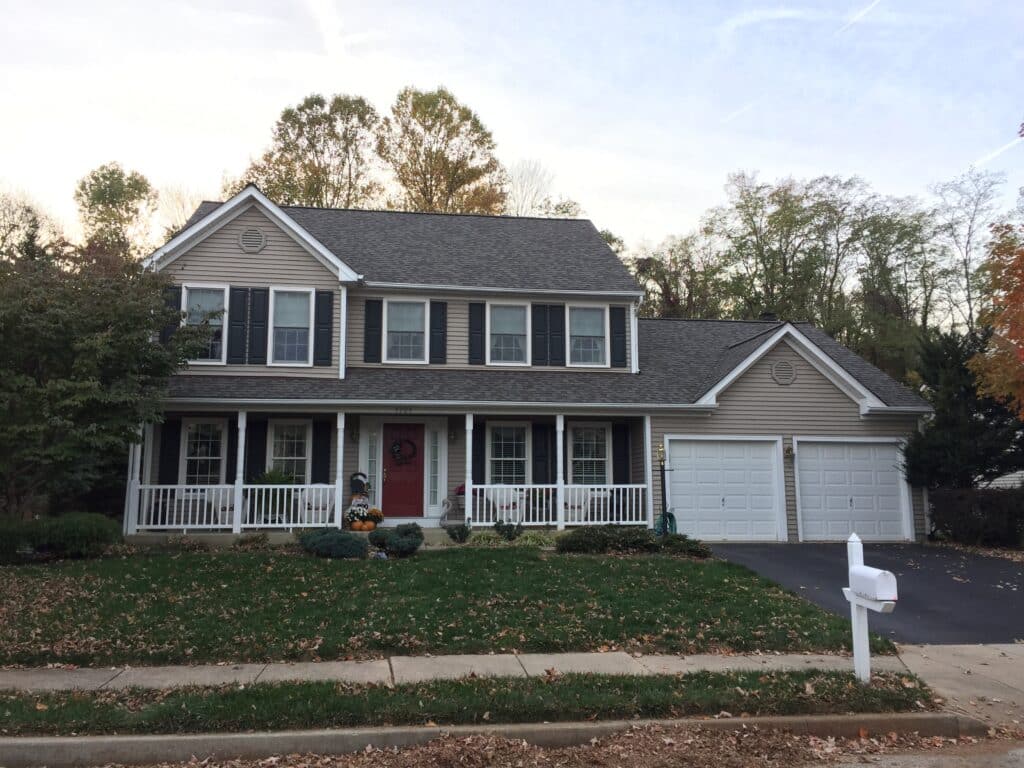 Front view of a beige two-story house with a white porch, red front door, and black shutters, pumpkins on the steps, and a two-car garage to the right.