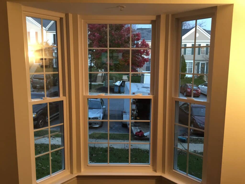 View from a bay window looking out onto a suburban street with red-leaved tree and parked cars across the curb at dusk