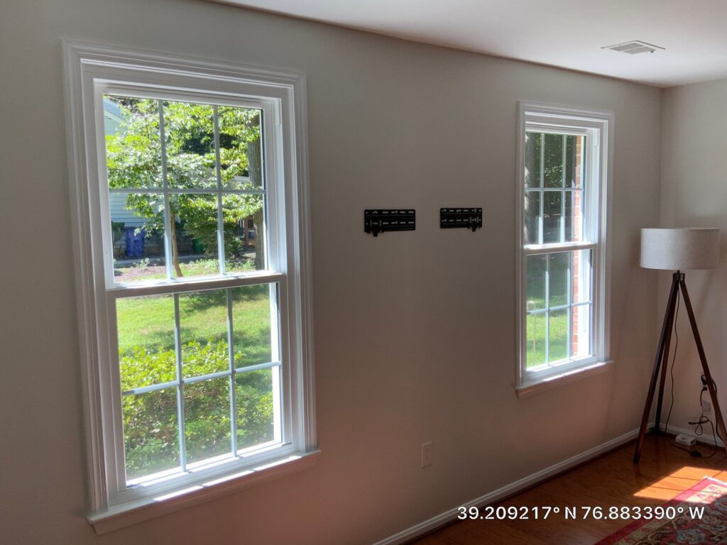 Two large white-framed windows in a bright beige living room, with sunlight on hardwood floor and a tripod lamp in the corner; coordinates overlay bottom right.