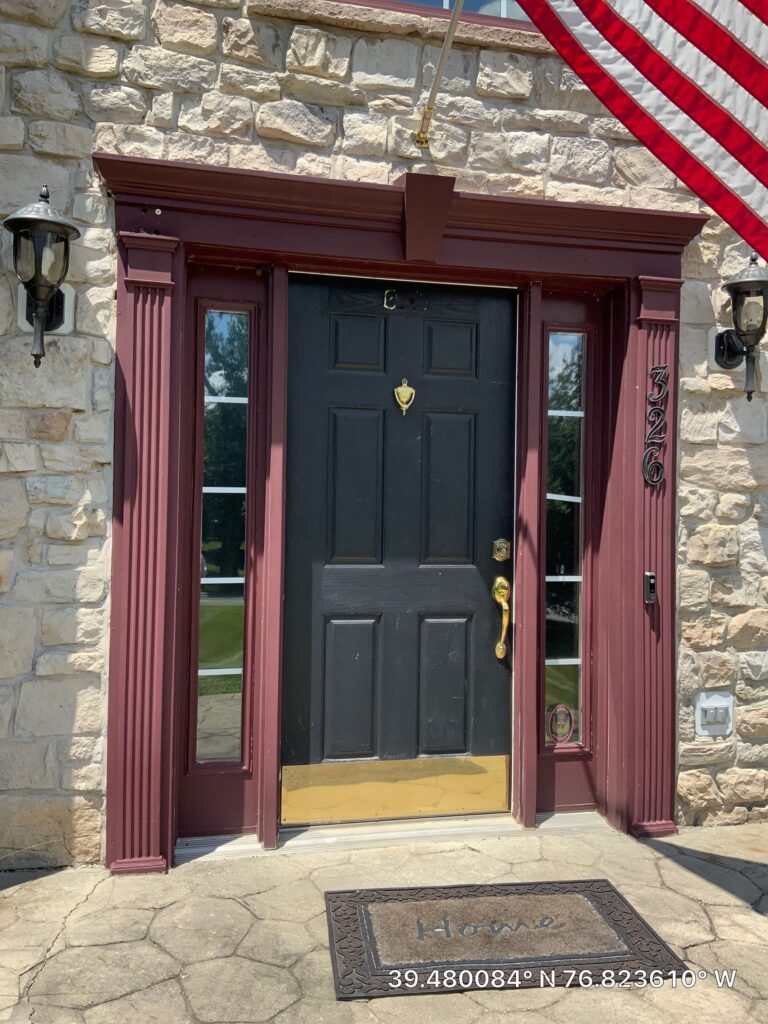 Stone house entry with burgundy trim around a black front door, brass knocker, and flag overhead on the right. Lantern lights flank the doorway.