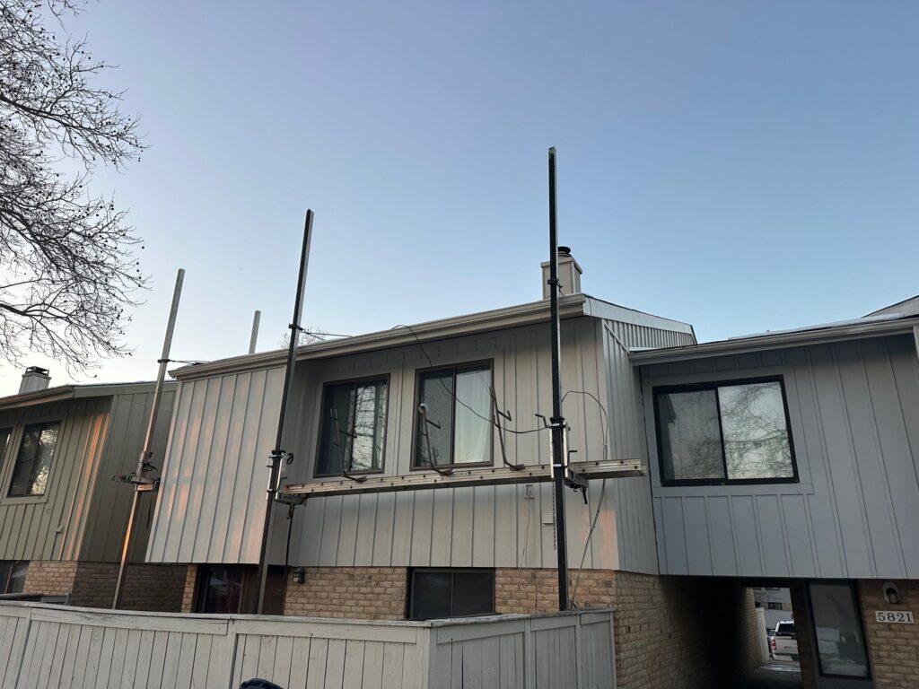 Two-story residential building with metal scaffolding poles outside, against a clear blue sky and a bare tree on the left.