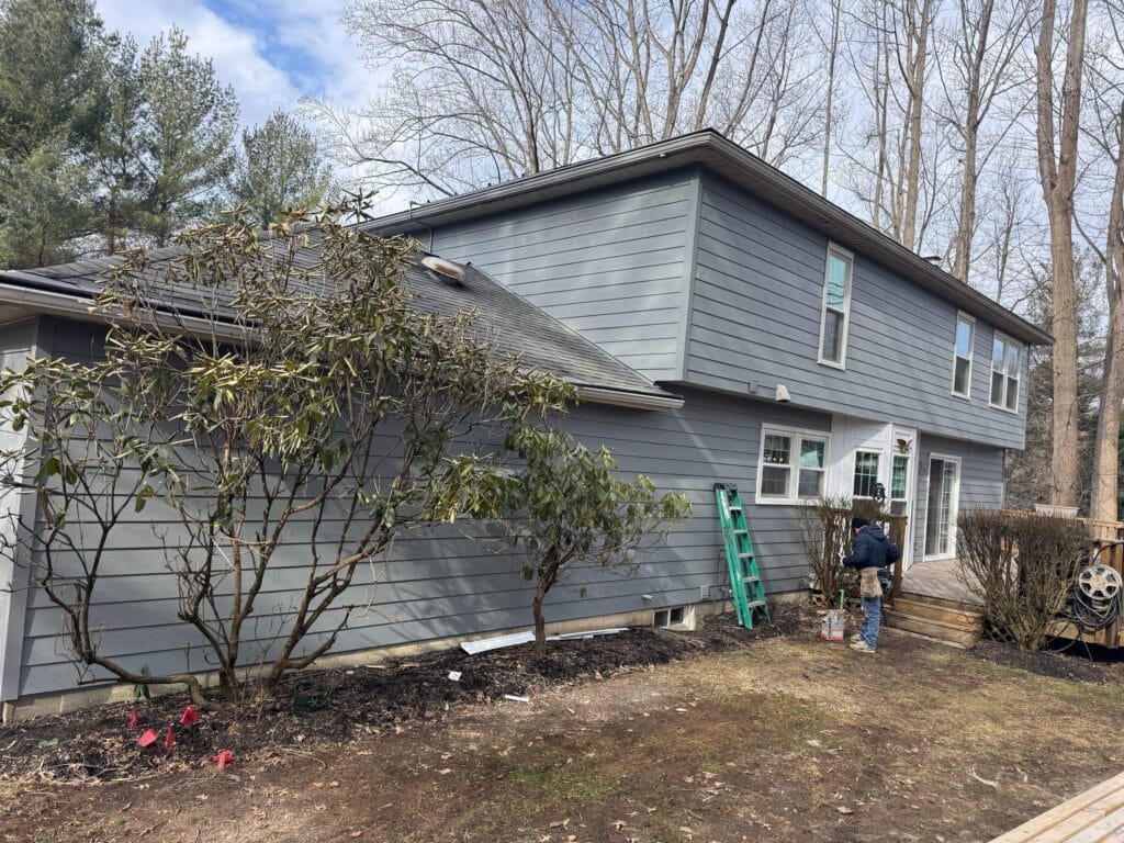 Two-story blue-gray house with a ladder propped against the siding and workers in the yard, bare trees in the background.