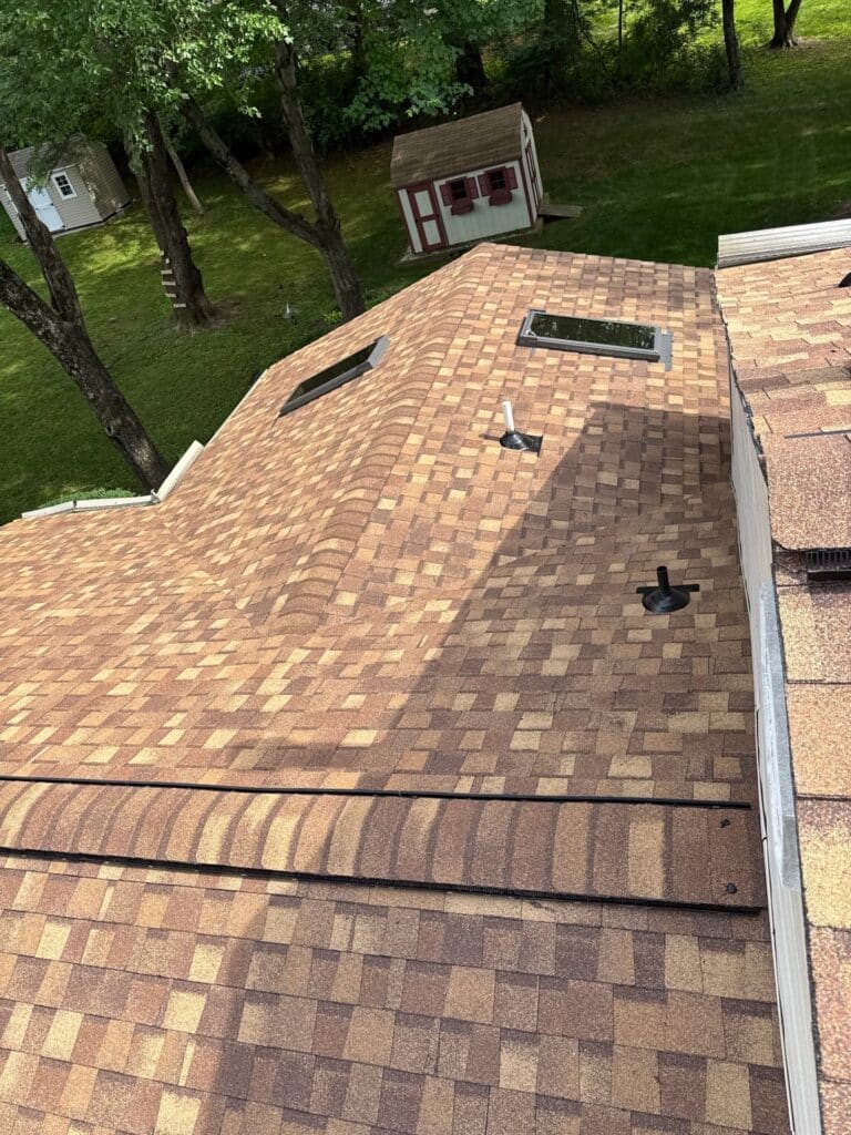 Aerial view of a brown shingled roof with skylights and vents, trees and a green lawn in the background and a small shed visible beyond the yard edge