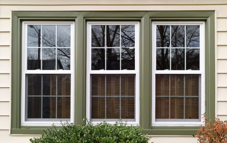 Three white-framed windows with green trim set in beige siding, reflecting trees and exposing blinds inside, with shrubs below.